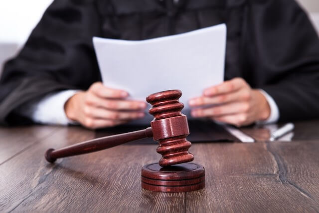 Wooden judge's gavel pictured on a desk in the foreground with a judge in a black robe reading a legal document in the background