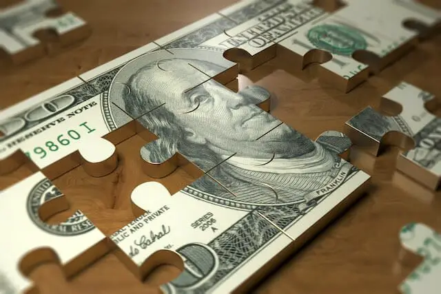 $100 bill on a wooden surface partially built out of puzzle pieces with the other pieces scattered around beside it