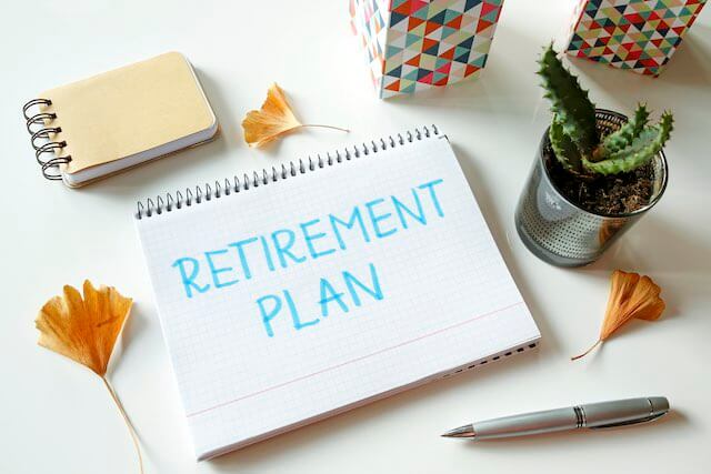 Words 'retirement plan' written on a notebook sitting on a desk next to a pen, notepad and decorative plant