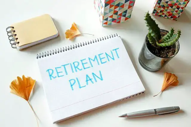 Words 'retirement plan' written on a notebook sitting on a desk next to a pen, notepad and decorative plant