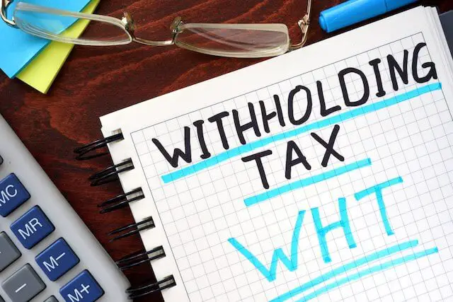 Words 'withholding tax' written on a note pad sitting on a desk next to glasses and a calculator