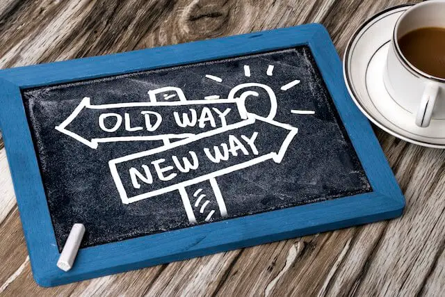 Chalkboard sitting on a wooden surface next to a coffee cup with two road signs drawn on it pointing in opposite directions, one which says 'old way' the other says 'new way' - change