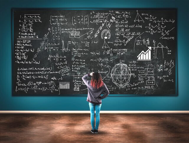 Young woman with her back to the camera scratching her head and appearing puzzled as she stares at a plethora of math problems drawn on a chalkboard