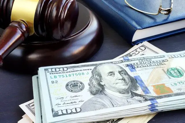 wooden judge's gavel on a desk next to a stack of $100 bills, a book and reading glasses