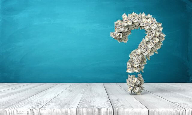 3D question mark made out of cash sitting on a wooden surface against a turquoise background - pay/money/salary questions