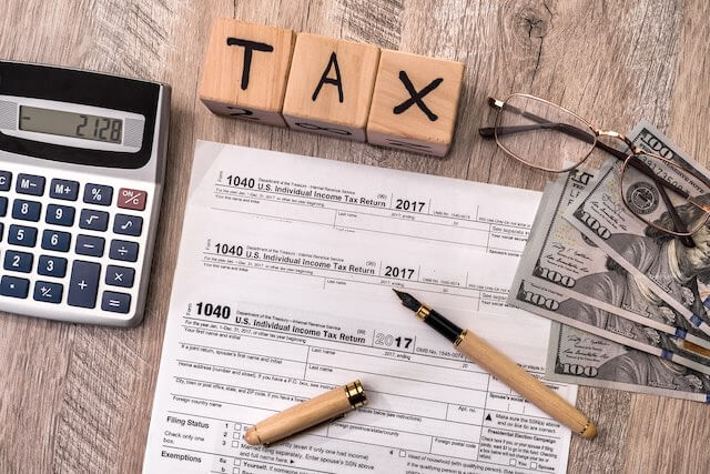 Words 'tax' spelled out by block letters above a spread of IRS 1040 income tax forms next to cash and a calculator on a desk
