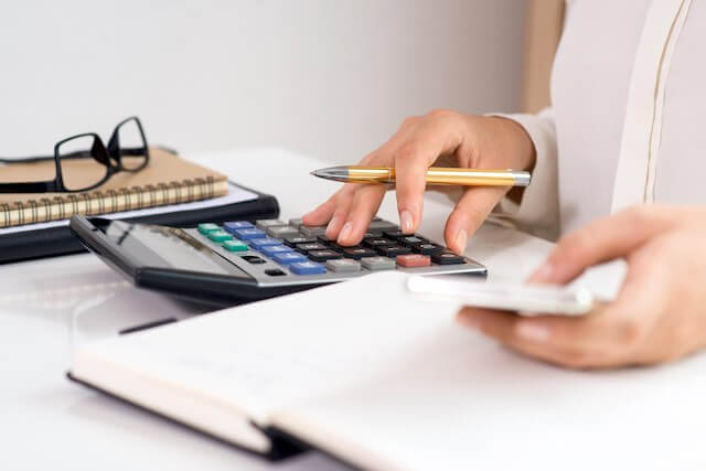 Close up of a woman's hands holding a smartphone and working on a calculator as she performs financial calculations while writing notes in a notebook