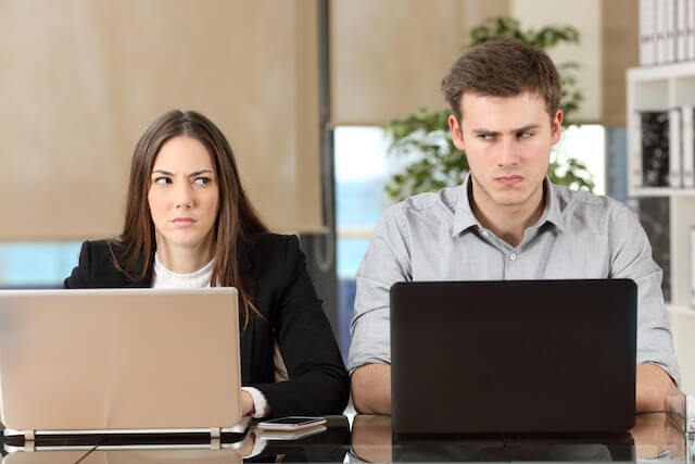 Male and female colleagues sitting next to each other working on their computers as they exchange angry glances at one another depicting frustration with co-workers, toxic working environment