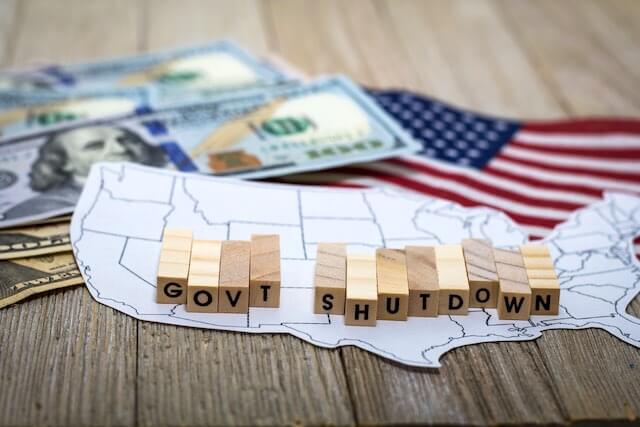 Wooden letters in a row that spell 'govt shutdown' on top of a map of the United States next to some cash and an American flag