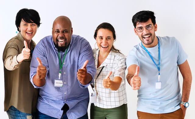 Group of 4 happy, smiling employees/co-workers standing in a row smiling and giving a thumbs up sign