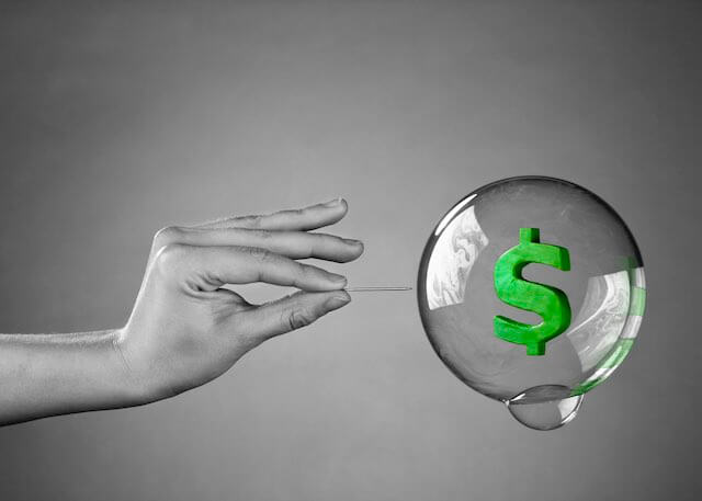 Close up of a person's hand holding a needle as it moves towards a bubble with a dollar sign inside of it depicting the bursting of an investment bubble