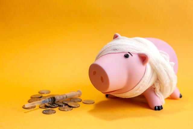 Pink piggy bank wrapped in gauze next to some coins and a syringe on a solid orange background depicting money/financial mistakes