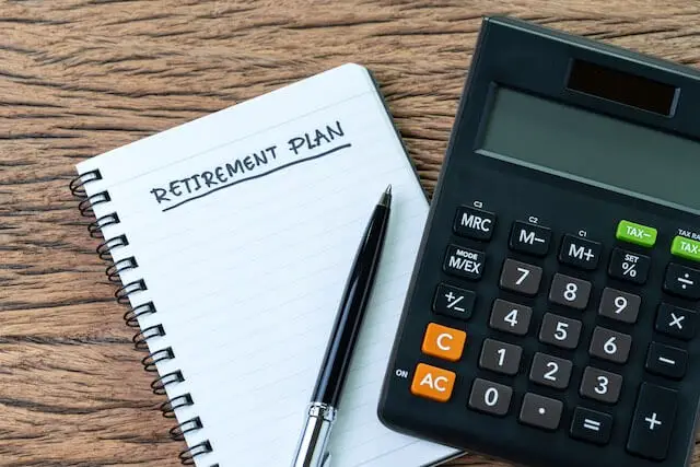 Words 'retirement plan' written at the top of a small notepad sitting on a wooden surface next to a pen and calculator