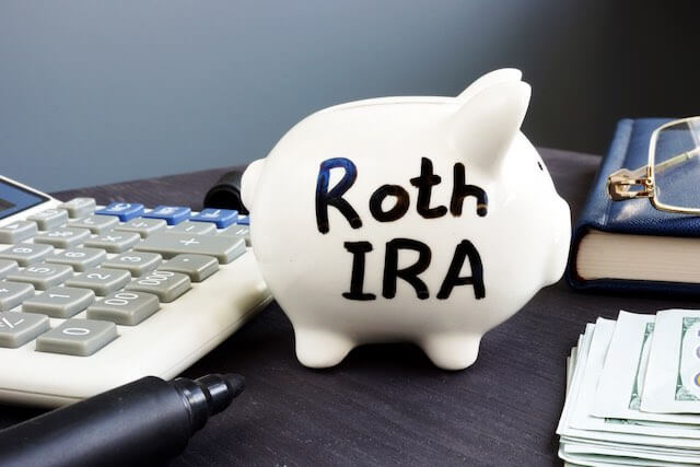 White piggy bank on a desk labeled 'Roth IRA" next to cash, a pen and a calculator - Roth TSP, retirement savings