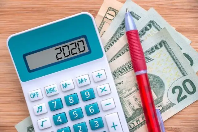 Calculator on a wooden surface with the numbers '2020' displayed on its face lying next to a pen and some cash