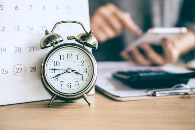 Old style alarm clock sitting on a desk in front of a desktop calendar and a blurred image of a businessman working on a calculator sitting at a desk in the background