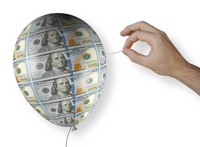 Close up of a person's hand holding a needle about to pop a balloon with images of $100 bills overlaid on it depicting a financial or stock market bubble