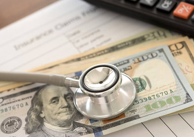 Stethoscope lying on top of a $100 bill next to a calculator and insurance paperwork depicting rising health insurance costs/premiums