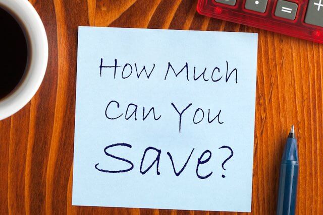 Words 'how much can you save?' written on a post-it note stuck to a wooden desk surface next to a coffee cup, pen and keyboard