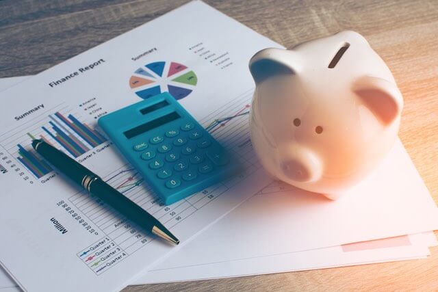 Piggy bank sitting on top of sheets of paper with financial graphs printed on them next to a calculator and pen