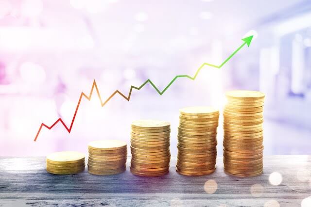 5 vertical stacks of coins growing in size from left to right with a line graph trend arrow pictured above them depicting rising costs or increasing investment returns