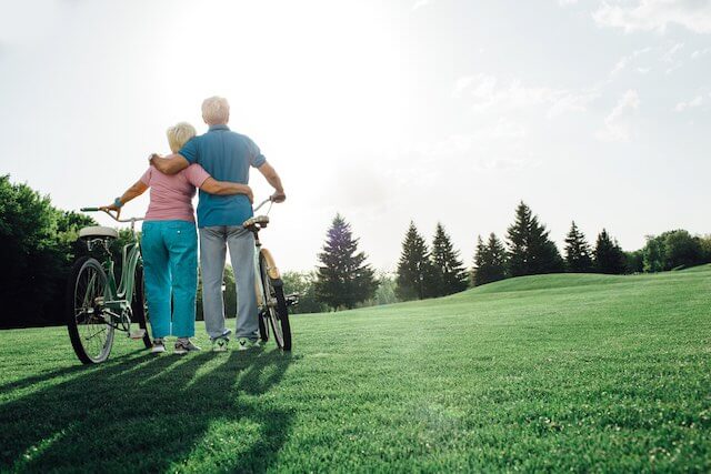 Senior couple with their backs to the camera standing embracing each other while they hold their bicycles beside them as they look out over a pretty green grass and hilltop scenic view