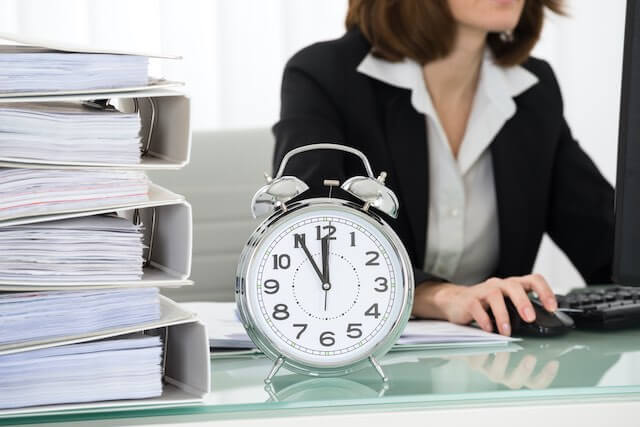 Old style alarm clock sitting on a desk next to a stack of binders with a businesswoman pictured working on a computer in the background