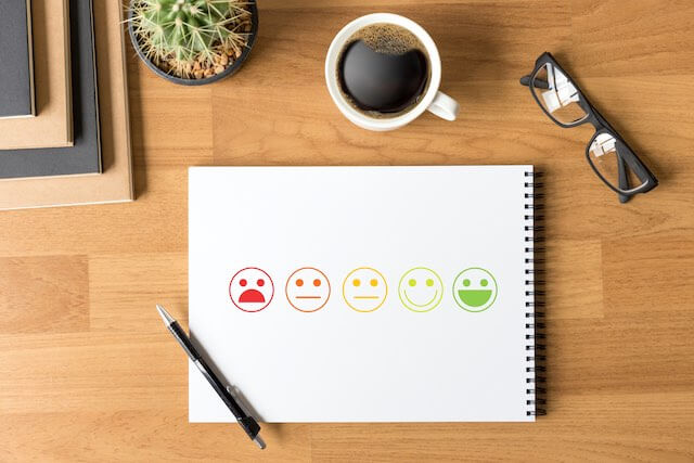 Notepad on a desk seen from above with drawings of emoji faces going from red and appearing upset to green and appearing very happy depicting a five point rating scale; notepad pictured next to glasses, coffee, and a small cactus at an employee's workstation