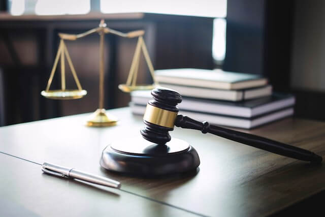 Wooden judge's gavel on a desk next to a pen, stack of law books, and justice scales