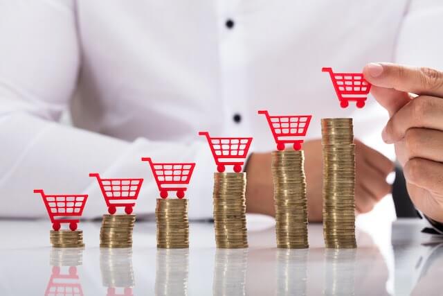 Stacks of coins growing in size from left to right with a closeup of a man's hand placing shopping cart replicas on top of each stack depicting rising inflation costs