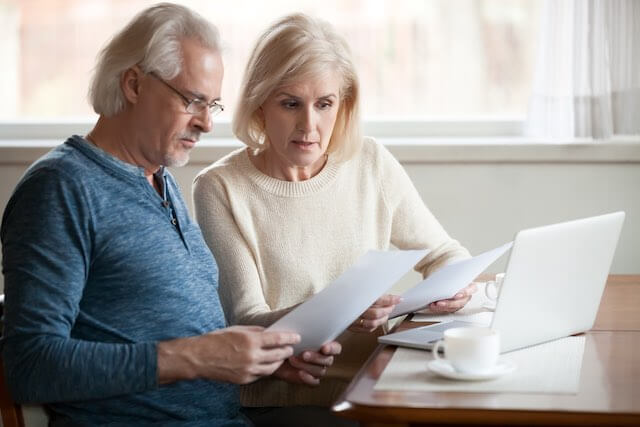 Senior aged couple sitting in front of a laptop at a desk as they carefully read and study some paperwork