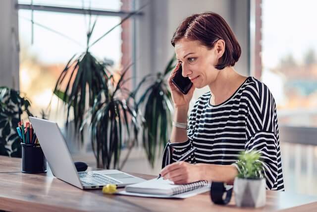 Woman talking on the phone and looking at a laptop as she teleworks from home