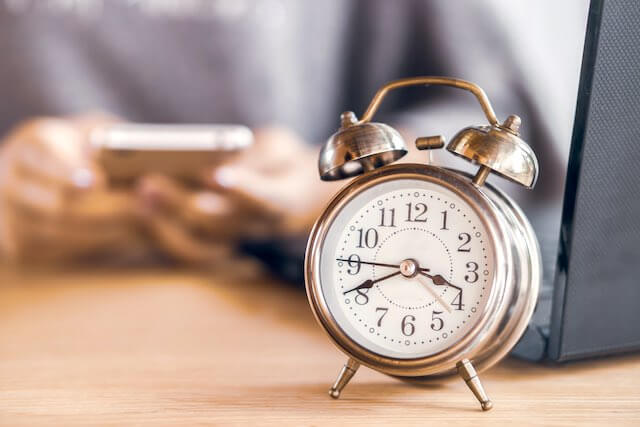 Alarm clock sitting on a desk with the blurred image of a person in the background in front of a laptop and working on a smartphone