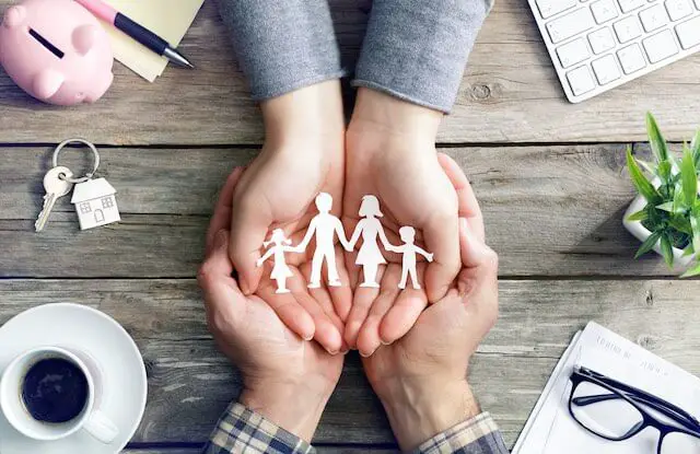 Close up of a man's and woman's hands cupped with palms facing upwards across a desk holding a paper cutout of a family (parents and two children)