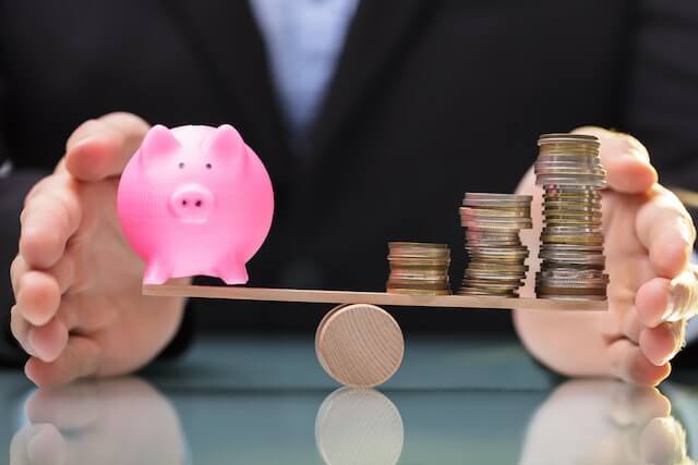 Man's hands on both sides of a seesaw as he works to balance the scale evenly between a ping piggy bank on one side and three stacks of coins growing in size vertically on the other