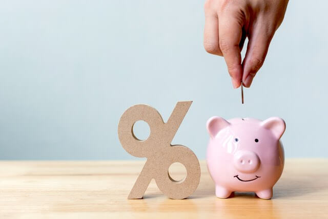 3D percent sign (%) sitting next to a pink piggy bank on a wooden surface with a close up of a person's hand putting a coin into the piggy bank