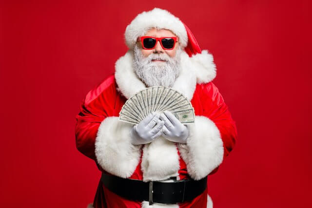 Santa Claus wearing beach sunglasses and holding a fan spread of cash in his hands against a solid red background