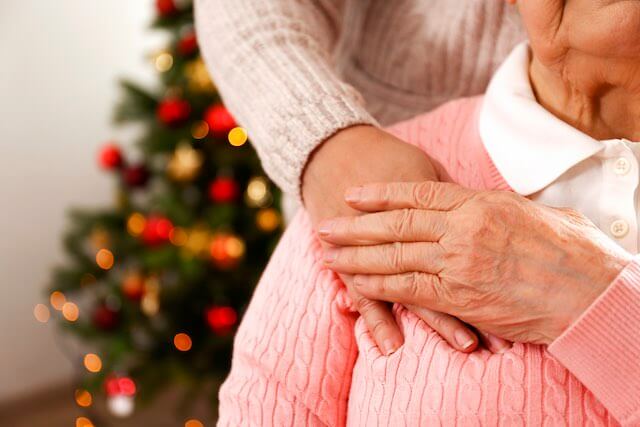 Close up of the shoulder of a senior citizen woman touching the hand of her daughter or caregiver standing behind her with a Christmas tree pictured in the background