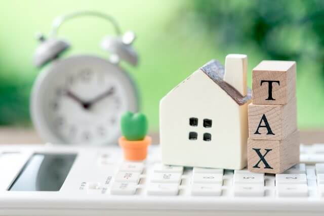 Small wooden model of a house next to block letters vertically stacked that spell 'tax' both sitting on top of a calculator with an old style alarm clock in the background