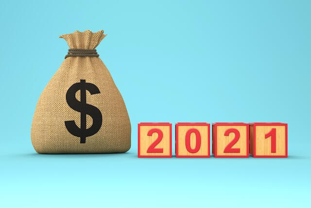 Bag of money next to wooden block numbers reading '2021' against a solid teal background