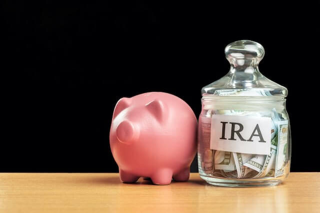 pink piggy bank next to a glass jar filled with cash labeled 'IRA' on a wooden surface