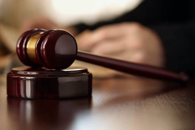 Wooden judge's gavel on a desk with a blurred image of a judge's hands in the background holding a legal brief