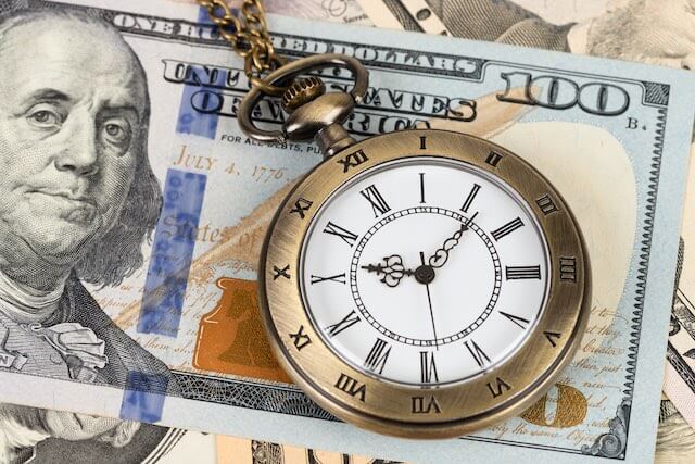 Close up of a $100 bill with a pocket watch on top of it depicting growth in the value of money over time