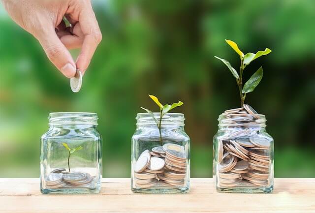 3 jars of coins side by side with a larger amount of coins in them going from left to right with green plants growing out of the top of the two larger ones; a close up of a man's hand puts money into the first jar as he adds to his retirement savings