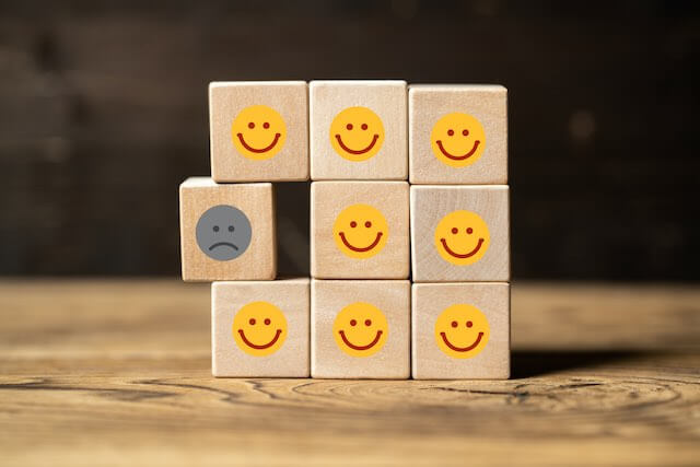 Square shaped stack of 9 wooden blocks, each continuing a smiling happy face, with one block jutting out slightly on the left side that contains an unhappy face depicting a single exclusion