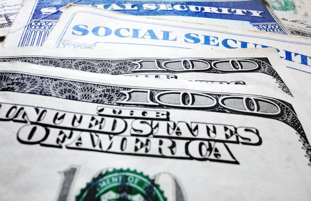 Close up of the words 'The United States of America' printed on a $100 bill in the foreground with another $100 bill underneath and two Social Security cards further back in the stack