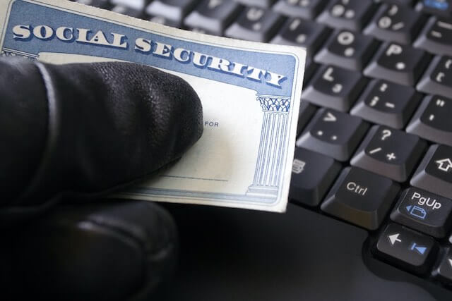 Close up of a person's hand wearing a black glove holding a Social Security card over a keyboard depicting fraud, scams or identity theft