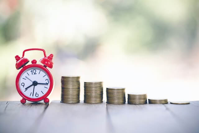 An old style red and white alarm clock sitting next to five vertical stacks of coins growing in size