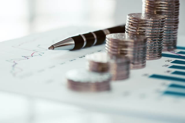 Vertical stacks of coins growing in size from one end to another on top of a printed page of bar graphs next to a pen