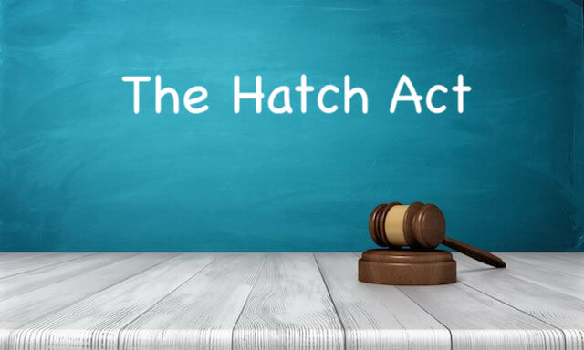 Wooden judge's gavel on a grey wooden surface with the words 'The Hatch Act' written on a teal chalkboard behind it